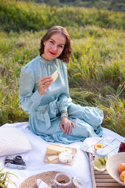 stock image A woman in a long summer dress with short hair sitting on a white blanket with fruits and pastries and eating cheese. Concept of having picnic in a city park during summer holidays or weekends. 