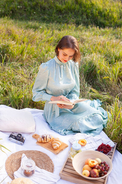 A woman in a long summer dress with short hair sitting on a white blanket with fruits and pastries and reading the book. Concept of having picnic in a city park during summer holidays or weekends. 