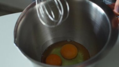 Hamur yapmak için yumurta çırpıyorsun. Eller, metal bir kâsede yumurta çırpar. Beyaz bir masada çırpar. Kapatın. El çırpıcısı, pişirmeden önce çırpılmış yumurta. Pasta yapma süreci.