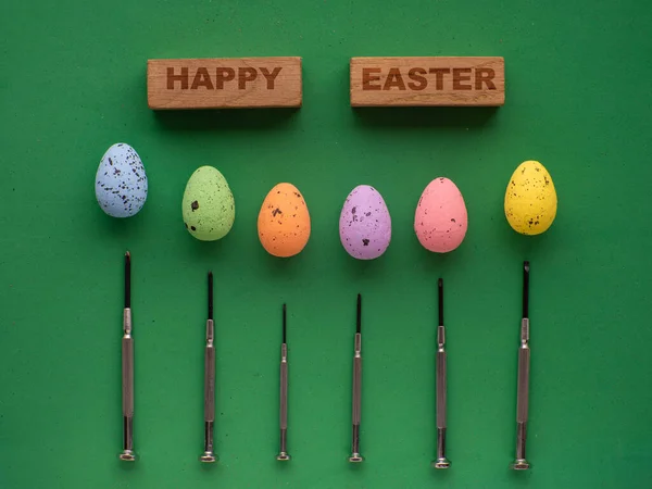 Easter eggs on green background with mini screwdrivers. Easter card design wih Happy Easter text on wooden blocks. Easter tools, worker concept.