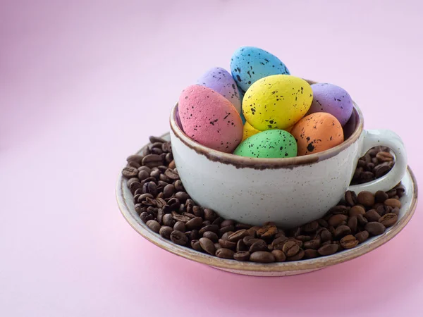 Composition with easter eggs in cup surrounded by coffee roasted beans. Easter coffee concept. Mug full of colorful eggs on a plate with coffee beans on a pink background with copy space.