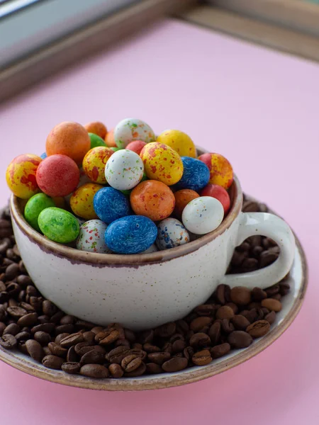 Composition with small chocolate easter eggs in cup surrounded by coffee roasted beans. Easter coffee concept. Mug full of colorful eggs on a plate with coffee beans on a pink background.