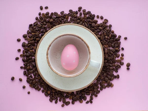 Flat lay composition with easter egg in cup surrounded by coffee roasted beans. Easter coffee concept. Mug full of pink egg on a plate on a pink background with a lot of coffee beans and copy space.