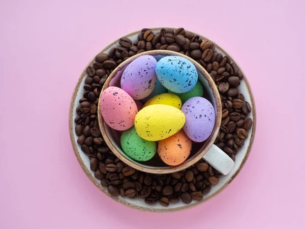 Flat lay composition with easter eggs in cup surrounded by coffee roasted beans. Easter coffee concept. Mug full of colorful eggs on a plate with coffee beans on a pink background with copy space.