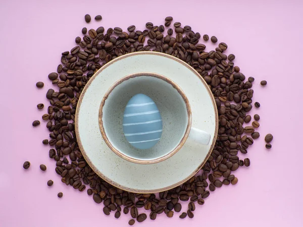 Flat lay composition with easter egg in cup surrounded by coffee roasted beans. Easter coffee concept. Mug full of gray egg on a plate on a pink background with a lot of coffee beans and copy space.
