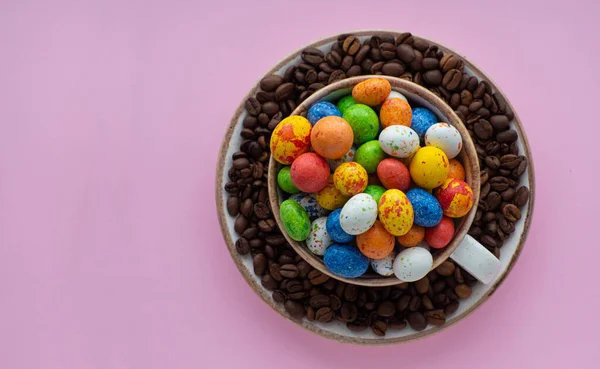 Flat lay composition with small chocolate easter eggs in cup surrounded by coffee roasted beans. Easter coffee concept. Mug full of colorful eggs on a plate with coffee beans on a pink background.