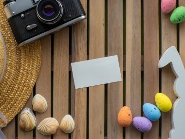 Hasır şapkalı ahşap arka plan, fotoğraf makinesi, kabuklar, renkli yumurtalar ve beyaz tavşan. Fotokopi alanı olan Paskalya kartı tasarımı. Paskalya ve seyahat konsepti. Tatillerde dinlenme ve seyahat.