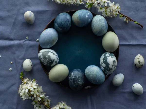Blue painted easter eggs. Dyed Easter eggs with marble stone effect blue color on blue tablecloth background with cherry blossom twigs. Eggs background with copy space, Ester holiday postcard concept.