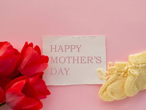 为新生儿准备的红色郁金香花和黄色靴 背景为粉色 母亲节快乐 母亲节贺卡 为母亲写祝福 用纸卡片和红花编织的婴儿鞋 — 图库照片