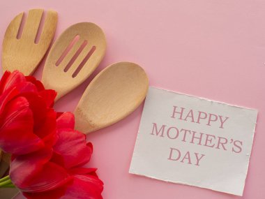 Kırmızı lale çiçekleri, mutfak aletleri ve pembe arka planda Mutlu Anneler Günü yazılı beyaz kağıt. Kadınlar ve Anneler Günü tebrik kartı. Doğum günü notu. Utensil buket düz yatıyordu.