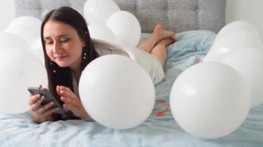 Woman in white clothes lying between white color balloons on a bed. Portrait of smiling woman using her phone with a lot of white air balloons. Happy birthday. Decoration for wedding. Copy space.