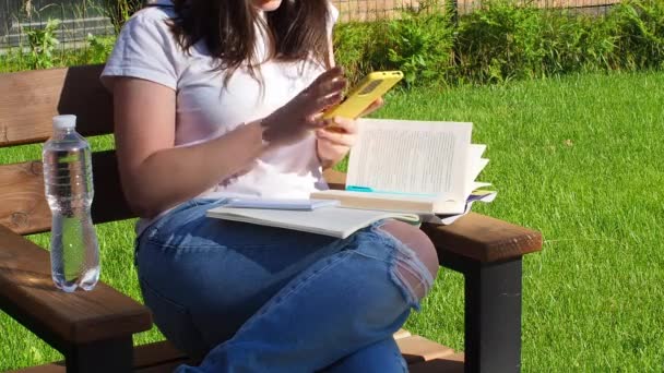 快乐的黑发女人坐在长椅上 在公园看书 教育学校和人的概念 在校园的长椅上学习的女生 女孩与书坐在一起 瓶子与水和写作 — 图库视频影像