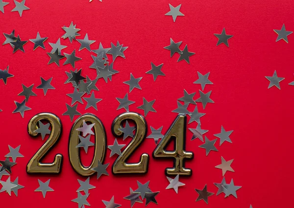 2024 on red background with silver stars and copy space. New year flat lay concept of greating card. Festive Christmas backdrop. Merry Christmas and Happy New Year. Silver star shaped confetti.