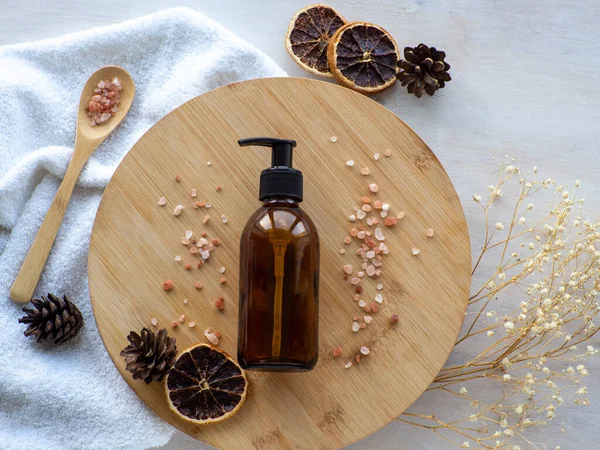 Natural winter skin care SPA beauty product design. Dark amber glass bottle on wooden plate with sea salt and cones. Mineral organic oil cosmetics on beige background. Christmas gift concept. Flat lay