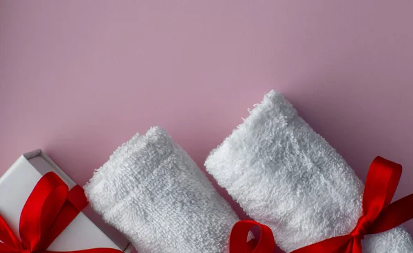 Flat lay with two white towels and gift box wrapped in white paper. Pink paper background with towels tied with a red bows. Valentines day spa, bath, self care and body care concept. Copy space.