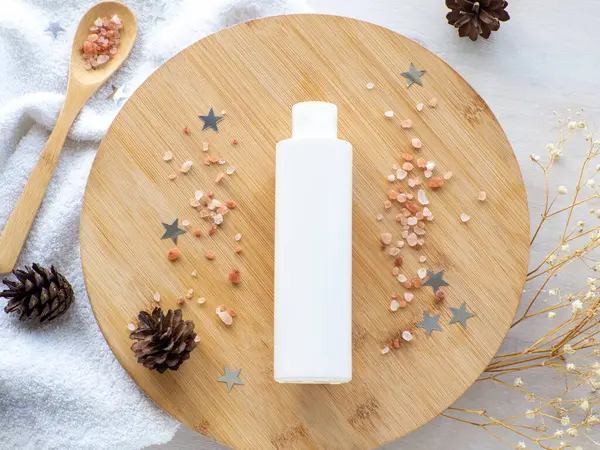 Natural winter skin care SPA beauty product design. White bottle on wooden plate with sea salt, stars and cones. Mineral organic oil cosmetics on beige background. Christmas gift concept. Flat lay