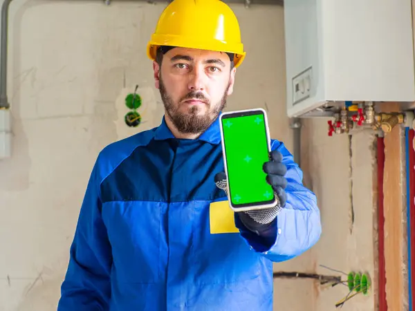 Portrait of worker or engineer in blue work clothes and yellow safety helmet with space for text. A man at a construction site holding a phone with chroma key screen in his hand and showing it.