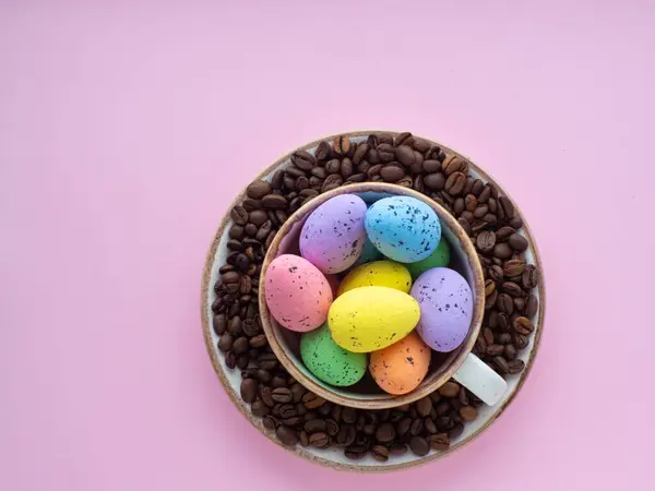 Flat lay composition with easter eggs in cup surrounded by coffee roasted beans. Easter coffee concept. Mug full of colorful eggs on a plate with coffee beans on a pink background with copy space.
