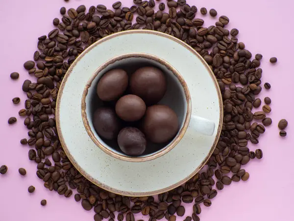 Flat lay composition with small chocolate easter eggs in cup surrounded by coffee roasted beans. Easter coffee concept. Mug full of sweet brown eggs on a plate with coffee beans on a pink background.