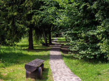 Bu görüntü, canlı yeşil bir parkın içinde kırsal ahşap bir bankın bulunduğu sakin bir taş yolu yakalıyor. Sahne ziyaretçileri dinlenmeye ve doğal güzelliğin tadını çıkarmaya davet ediyor..