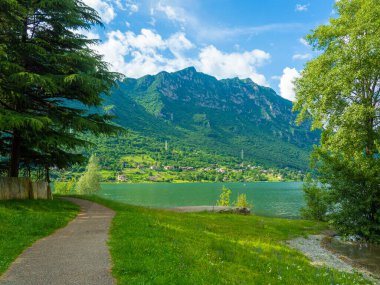 Bulutlu gökyüzünü yansıtan sakin bir göl içeren çarpıcı bir manzara. Yeşillik tarafından çerçevelenmiş görkemli dağlar uzaklardan yükselirler, sakin ve resimsel bir manzara yaratırlar. İdro Gölü, Kocakarı, İtalya