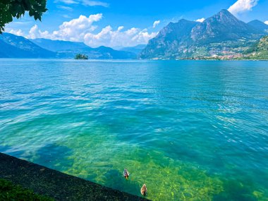İtalya 'daki mavi gökyüzünü yansıtan sakin bir Iseo gölünün olduğu çarpıcı bir manzara. Yeşillik tarafından çerçevelenmiş görkemli dağlar uzaklardan yükselir. Huzurlu ve görkemli bir manzara yaratır..