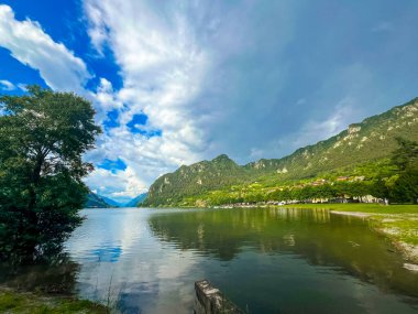 Bulutlu gökyüzünü yansıtan sakin bir göl içeren çarpıcı bir manzara. Yeşillik tarafından çerçevelenmiş görkemli dağlar uzaklardan yükselirler, sakin ve resimsel bir manzara yaratırlar. İdro Gölü, Kocakarı, İtalya