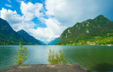 Bulutlu gökyüzünü yansıtan sakin bir göl içeren çarpıcı bir manzara. Yeşillik tarafından çerçevelenmiş görkemli dağlar uzaklardan yükselirler, sakin ve resimsel bir manzara yaratırlar. İdro Gölü, Kocakarı, İtalya