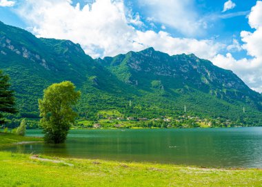 Bulutlu gökyüzünü yansıtan sakin bir göl içeren çarpıcı bir manzara. Yeşillik tarafından çerçevelenmiş görkemli dağlar uzaklardan yükselirler, sakin ve resimsel bir manzara yaratırlar. İdro Gölü, Kocakarı, İtalya