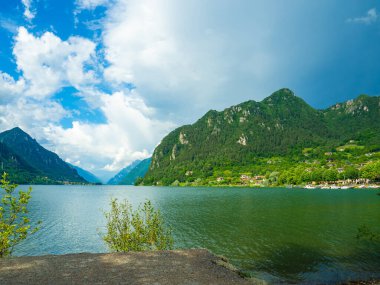 Bulutlu gökyüzünü yansıtan sakin bir göl içeren çarpıcı bir manzara. Yeşillik tarafından çerçevelenmiş görkemli dağlar uzaklardan yükselirler, sakin ve resimsel bir manzara yaratırlar. İdro Gölü, Kocakarı, İtalya