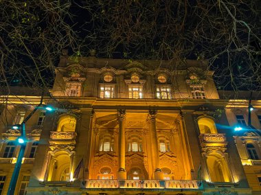 Budapeşte Üniversitesi binasının dış cephesinde ağaçlarla çevrili sarı ve mavi ışıklar olan bir gece çekimi..