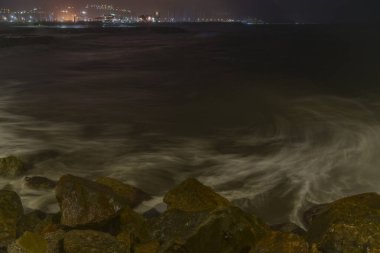 Güzel bulanık deniz dalgaları kumlu ve kayalık bir sahilde, arka planda aydınlık bir şehir