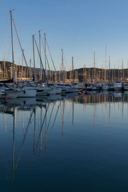 Sakin bir limanda yelkenli ve yatları olan bir liman, güneşle güzelce aydınlatılmış.