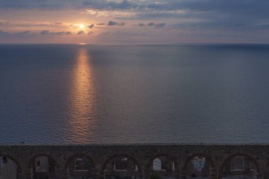 Sahilde gün batımının manzarası. Önünde terk edilmiş eski bir bina ve sakin bir su yüzeyi var.