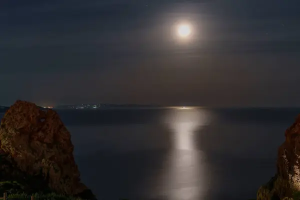 Deniz üzerinde dolunay manzarası. Arka planda şehir ve ön planda kayalar var..