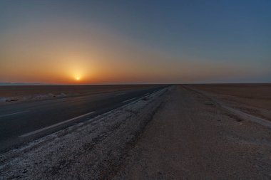 Gün batımında çölün ortasından ufka uzanan asfalt bir yol manzarası.