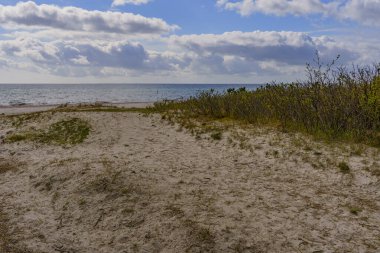 Açık denizin geniş manzarası, kumlu bir sahilden görülebiliyor. Gökyüzü çimlerle kaplı ve arka planda bulutlar.