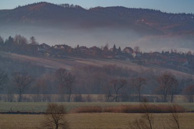 Şafak vakti tepelik bir arazide inşa edilmiş küçük bir konutun puslu manzarası.