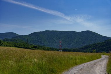 Uzak dağları ve kırmızı-beyaz yüksek voltajlı kulesi olan bir çakıllı yol çayırlardan geçer..