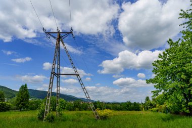 Yeşil bir çayırda duran üç ayaklı beton bir elektrik direği. Uzak dağlar, ağaçlar ve beyaz bulutlarla dolu mavi bir gökyüzü..