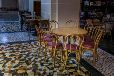 Round wooden table with classic cafe chairs stands on patterned tiles in an open-air seating area at dusk.