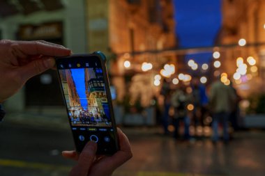 Close-up of hands framing a lively evening street scene on a phone screen, with glowing lights softly blurred.