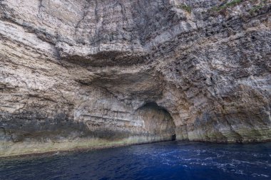 Derin mavi su ve köpüklü dalgaların üzerinde kemerli mağaranın açıldığı desenli uçurum duvarı yükseliyor..