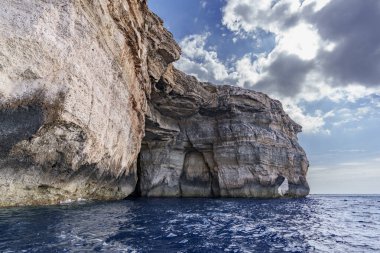 Büyük kireçtaşı burnu, mağara açıklıkları, dramatik güneş ışığı bulutlarının altında dalgalı mavi suyun üstünde kuleler..