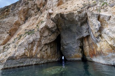 Sheer rock face reveals narrow twin cave openings above emerald water, textured strata and rugged formations.