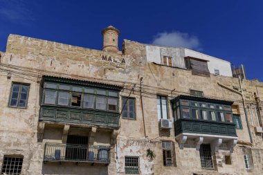Weathered historic building featuring enclosed timber balconies, peeling textures, chimney and blue Mediterranean sky.