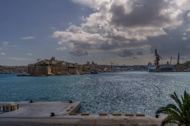 Wide harbor view with medieval fort, industrial shipyard, cruise ship and shimmering sea under dramatic clouds.