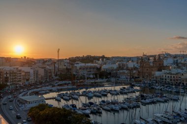 Warm golden sun sets behind city, illuminating yacht-filled marina and domed church on tranquil water.