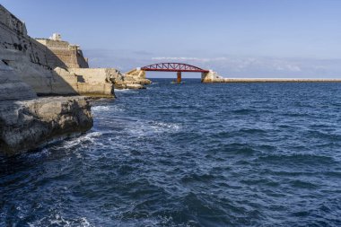 Stone fortifications and vivid red bridge facing dynamic blue waves along scenic Maltese coastline.