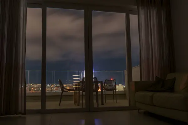 Modern living room opens to balcony with chairs and table, overlooking illuminated city under clouds.